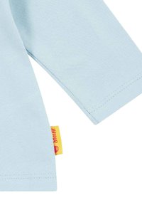 Helles blaues Langarmhemd mit sichtbaren Nähten und einem kleinen gelben Stoffetikett mit rotem Text an der Seitennaht.