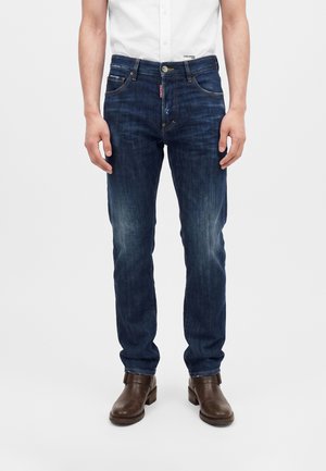 Homme portant un jean bleu foncé délavé, une chemise blanche à manches courtes et des bottines en cuir marron, debout devant un fond uni.