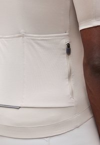 Κοντινό πλάνο μια τσέπης μπεζ υφασμάτινης φανέλας ποδηλασίας με φερμουάρ στο πλάι, φορεμένη από άτομο με σκούρο δέρμα και κοντό μανίκι.