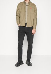 Camicia verde oliva con bottoni e maniche lunghe, abbinata a jeans neri e scarpe da ginnastica nere, presenta una tasca frontale e un logo patch.