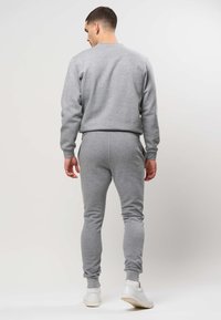 Mees seisab seljaga kaamera poole, kandes halli pusa, sobivaid trennipükse ja valgeid tossusid heledal ühtlasel taustal.