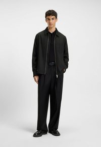 Sort lynlåsjakke med en lomme, lagdelt over en sort poloshirt, kombineret med løst siddende sorte bukser og sorte sko. Glat tekstur.