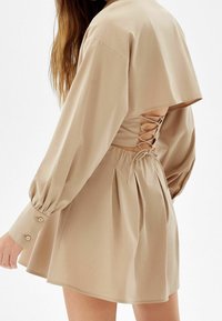 Beige klänning med avslappnad passform, långa puffärmar och snörning i ryggen. Tyget är mjukt och har insydda veck vid midjan.