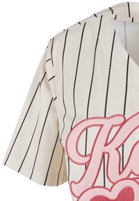 Baseball-inspirert jersey i kremfarge med svarte vertikale striper. Har et rosa brodert logo og hjerte-design på forsiden. Korte ermer.