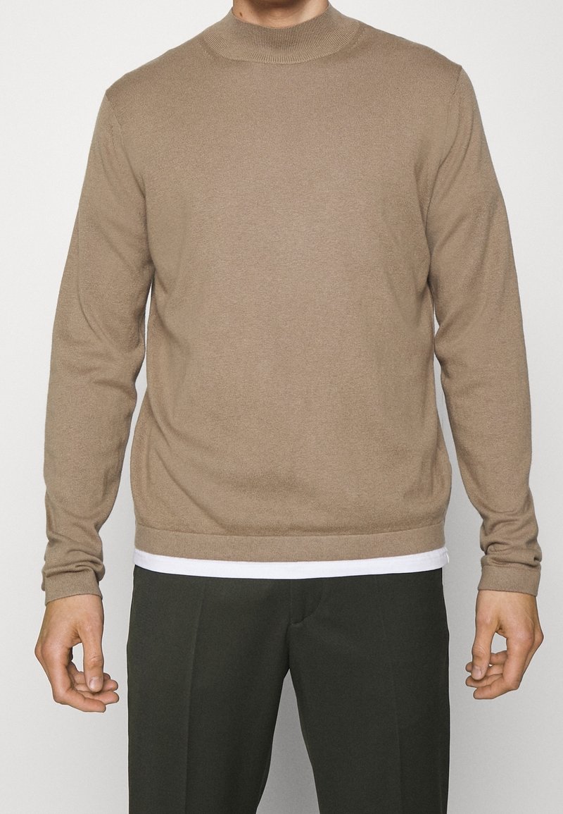 Homme portant un pull beige à manches longues sur une chemise blanche avec un pantalon vert foncé, debout devant un fond clair uni.