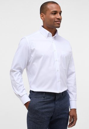 Chemise blanche à manches longues avec un col et une poche poitrine, associée à un pantalon texturé foncé. Tissu lisse et coupe ajustée.