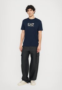 Tamsiai mėlynas medvilninis marškinėlis su auksiniu "EA7 EMPORIO ARMANI" grafiniu dizainu, derinamas su juodomis cargo kelnėmis su šoninėmis kišenėmis ir laisvu siluetu.