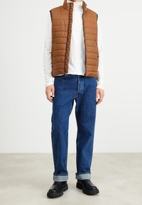 Braune, gesteppte Weste mit hohem Kragen, schwarze Seitenteile; getragen über einem weißen Hemd und blauen Jeans; schwarze Stiefel mit robuster Sohle.