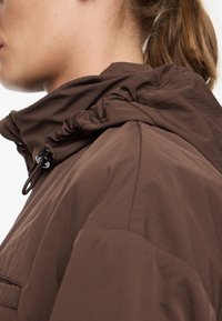 Chaqueta ligera marrón con un cuello bajo. Cuenta con un ajuste elástico alrededor del cuello y un bolsillo lateral. Textura suave con un diseño minimalista.