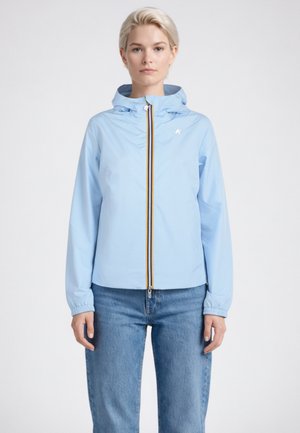 Donna che indossa una giacca con cappuccio azzurra chiusa con una zip, abbinata a jeans blu, in piedi di fronte a uno sfondo chiaro e uniforme.