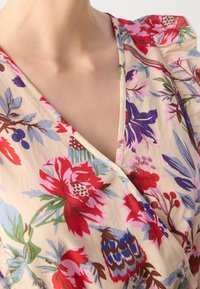 Gros plan d'une personne portant un chemisier beige avec un motif floral coloré en rouge, violet, bleu et vert, avec un col en V.
