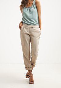 Top in satin azzurro chiaro con scollatura a volant, abbinato a pantaloni beige su misura arrotolati sui polsini e sandali neri con cinturini.