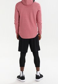 Rotes Sweatshirt mit Kapuze, kombiniert mit schwarzen Shorts und Leggings. Der Stoff sieht weich aus und hat eine leicht strukturierte Oberfläche. Sportliche Schuhe vervollständigen das Outfit.