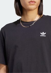Camiseta de algodón negra con cuello redondo, mangas cortas y un logotipo bordado de Adidas en blanco en el lado izquierdo del pecho. Textura suave.