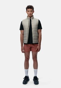 Lys grå puffer vest, uten ermer, med høy krage. Bært over en svart skjorte og kombinert med rustfargede shorts, svarte sko og hvite sokker.