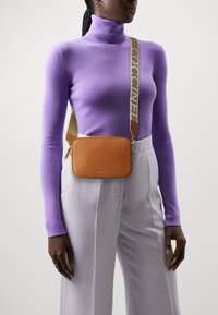 Braon kožni crossbody torba s teksturom i kontrastnim krem trakama na kojima stoji natpis "Coccinelle" u podebljanom slovima, nošena preko ljubičastog rolka.