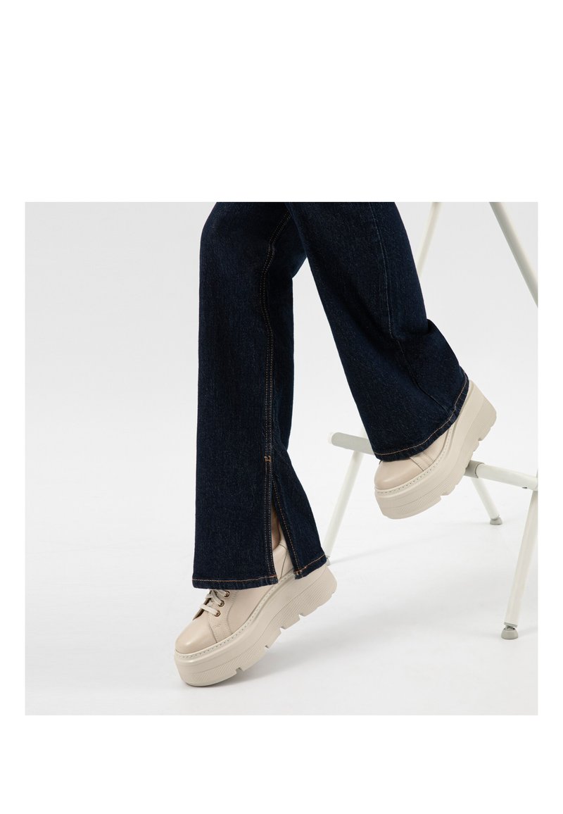 Osoba oblečená v tmavě modrých džínách s bočními rozparky a krémových botách na platformě se šněrováním, sedící na bílém skládacím křesle.