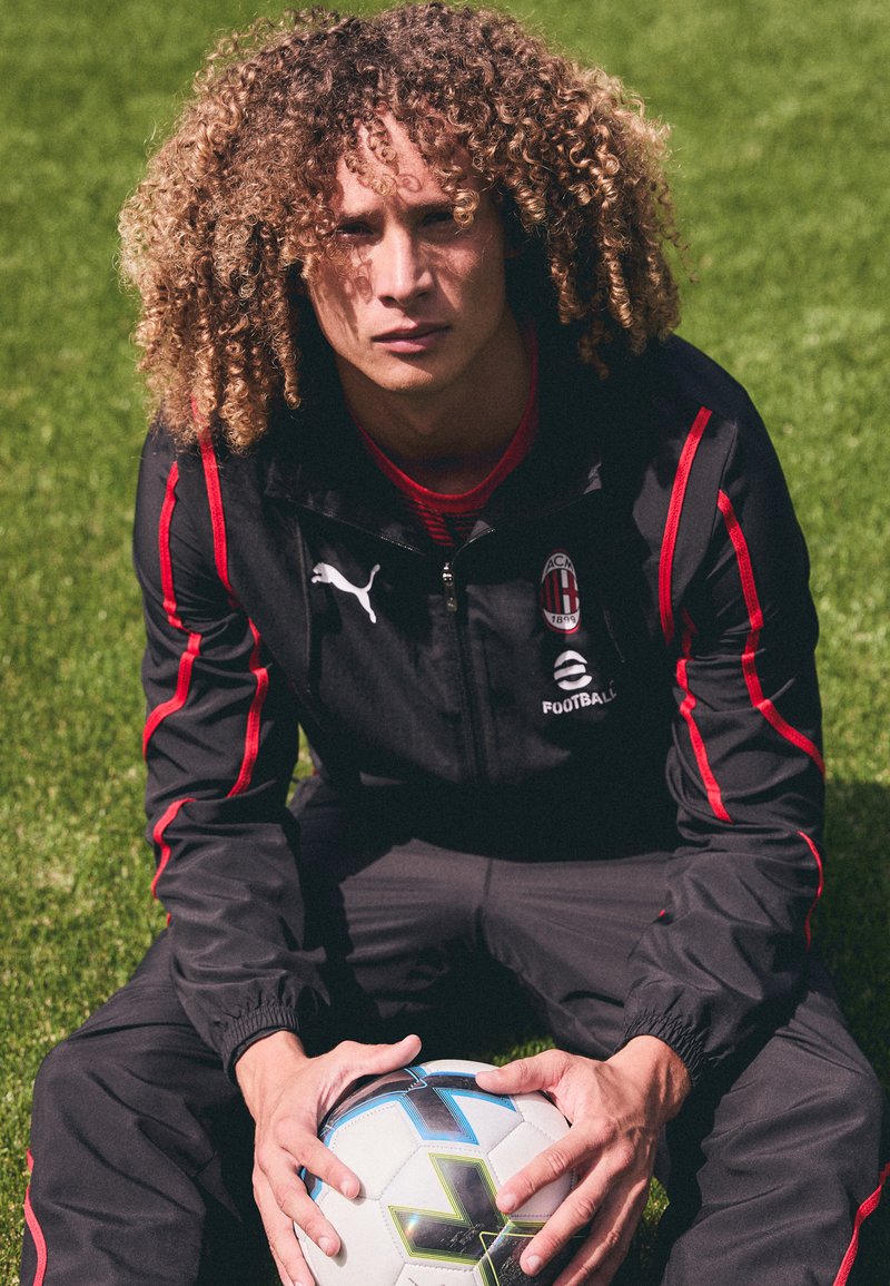 Chaqueta deportiva negra con rayas rojas y logo, acompañada de pantalones a juego. La persona sostiene un balón de fútbol en blanco y negro sobre hierba verde.