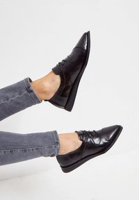 Chaussures oxford en cuir verni noir avec un bout pointu, un laçage, et un subtil détail perforé sur le dessus. Semelle en caoutchouc plate.