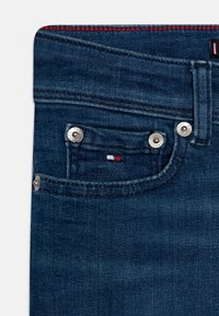 Jeans de mezclilla azul oscuro, con herrajes plateados, costuras rojas y un pequeño logotipo bordado con acentos blancos y rojos en el bolsillo.
