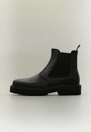 Botte Chelsea en cuir noir avec panneaux latéraux élastiques, languette au talon et semelle épaisse à crampons sur un fond clair uni.