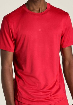 Uomo che indossa una semplice maglietta sportiva rossa a maniche corte con un piccolo e discreto logo al centro del petto.