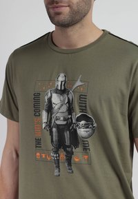 Hombre con camiseta verde que tiene una ilustración de Mandalorian y Grogu, con el texto "El niño viene conmigo" y símbolos de Star Wars en naranja y negro.