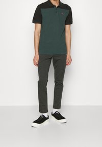 Polo tričko v tmavě zelené barvě s černým límečkem, krátké rukávy a logo. V kombinaci se slim-fit šedými kalhotami a černými teniskami s bílou podrážkou.