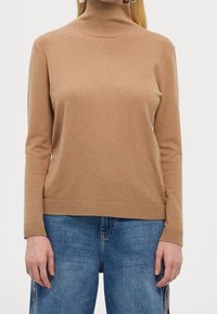 Pull en col roulé beige à manches longues, avec des poignets et un ourlet côtelés, fabriqué dans un matériau en tricot lisse, associé à un jean en denim bleu.