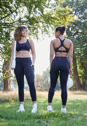 Sutiãs desportivos e leggings pretos em duas modelos, apresentando um design de costas nadador, tecido elástico e ténis brancos em contraste, situados numa área gramada.