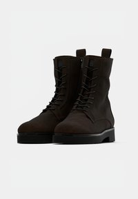 Bottines en suède marron avec un bout rond, un design à lacets à l'avant, une fermeture éclair sur le côté et une semelle en caoutchouc noir épaisse. Esthétique propre et minimaliste.