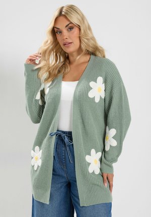 Lyshåret kvinde iført en grøn strikket cardigan med store hvide og gule tusindfrydsmønstre over en hvid top og blå, højtaljede jeans.