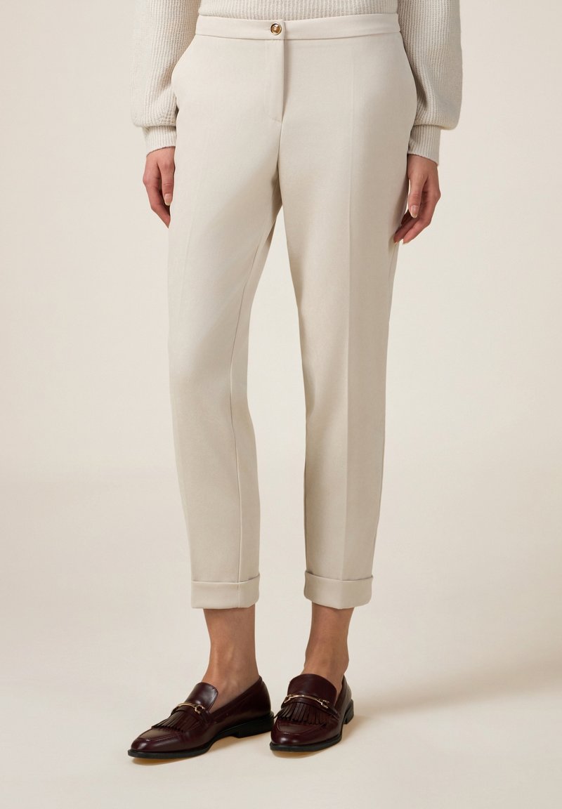 Pantaloni beige su misura con una texture liscia, orli risvoltabili e chiusura con bottone. Abbinati a mocassini marrone scuro con dettaglio frangia.