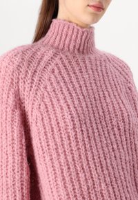 Rosa gestrickter Pullover mit hohem, geribbtem Kragen, der ein dickes, strukturiertes Maschenmuster und überschnittene Schultern aufweist.