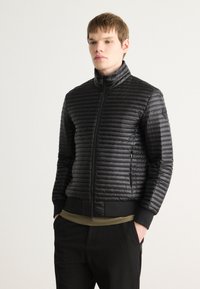 Melnā pufotā jaka ar augstu apkakli, horizontālu quilted dizainu, ar rāvējslēdzēju aizdari un elastīgām aprocēm. Uzvilkta virs olīvzaļas krekla.