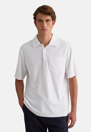 Jeune homme aux cheveux bruns portant un polo blanc à manches courtes avec une poche poitrine et un pantalon bleu marine, debout avec une main dans la poche.