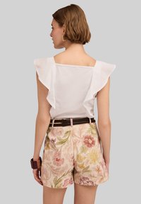 Weißes Oberteil mit Rüschensleeves, kombiniert mit hellrosa Blumenshorts. Brauner Gürtel und strukturiertes Blumenmuster auf den Shorts, vor einem grauen Hintergrund.
