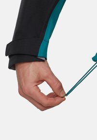 Zwarte en teal jas manchet met verstelbare elastische toggle. Het materiaal lijkt lichtgewicht met een gestructureerde afwerking. Hand grijpt de togglekoord.