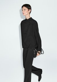 Outfit nero composto da una maglietta drappeggiata con collo alto e maniche ampie, abbinata a pantaloni aderenti e una piccola borsa nera.