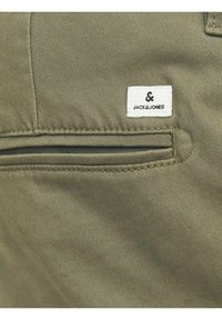 Gros plan d'un pantalon en tissu kaki avec une poche passepoilée à l'arrière et une étiquette blanche rectangulaire avec le logo Jack & Jones au-dessus de la poche.