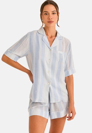 Vrouw draagt een lichtblauw-wit gestreept overhemd met korte mouwen en bijpassende shorts, staat met één hand op haar heup.