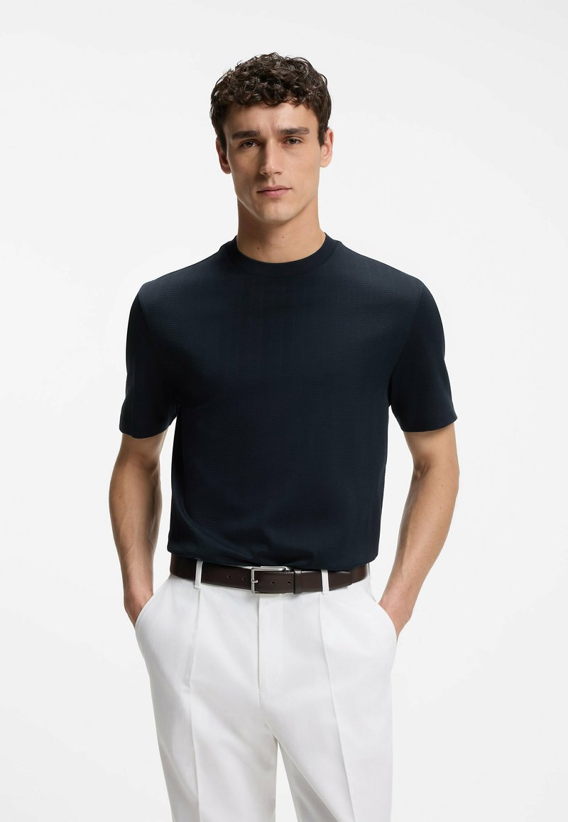 Jeune homme portant une chemise noire ajustée à manches courtes rentrée dans un pantalon blanc avec une ceinture marron, debout les mains dans les poches.