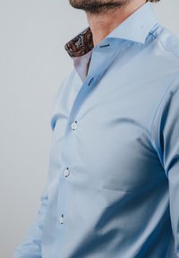 Man die een lichtblauw overhemd draagt met witte knopen en een patroon aan de binnenkant van de kraag, afgebeeld van nek tot taille tegen een effen achtergrond.