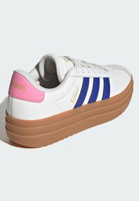 Białe trampki z trzema niebieskimi paskami, różowym akcentem na pięcie oraz teksturowaną brązową podeszwą na platformie. Na boku znajduje się złote logo.