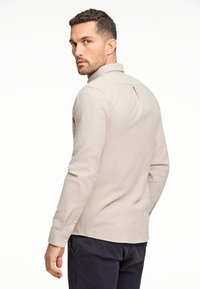 Camicia a maniche lunghe di colore beige chiaro con colletto, polsini con bottoni e dettaglio di piega posteriore. Il tessuto ha un aspetto morbido e testurizzato.