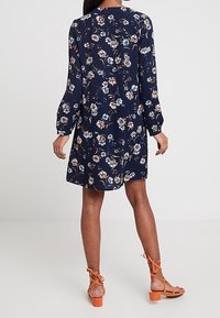 Abito floreale blu navy con maniche lunghe, caratterizzato da un motivo floreale più chiaro e una vestibilità comoda. Abbinato a sandali arancioni con cinturini.