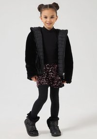 Giacca imbottita nera sopra una maglietta nera, abbinata a una gonna nera con paillettes luccicanti e leggings neri texturizzati. Completato con stivali neri foderati in pelliccia.