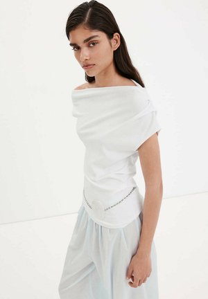 Femme aux longs cheveux foncés porte un haut blanc à épaules dénudées avec une ceinture chaîne et un pantalon ample bleu clair sur fond blanc uni.