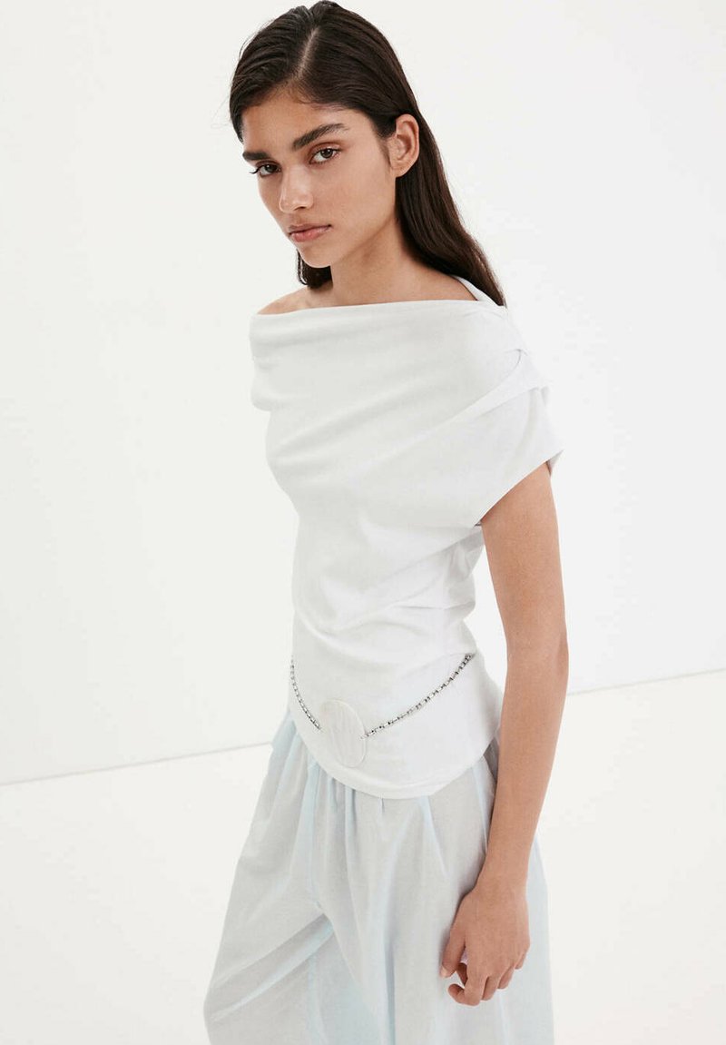 Femme aux longs cheveux foncés porte un haut blanc à épaules dénudées avec une ceinture chaîne et un pantalon ample bleu clair sur fond blanc uni.
