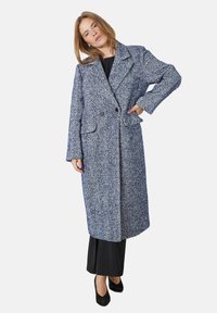Dolga modra in bela plašča s houndstooth vzorcem, s označenim ovratnikom, dvojno gumbom, stranskimi žepi in ohlapnim krojem, nad črnim kompletom.